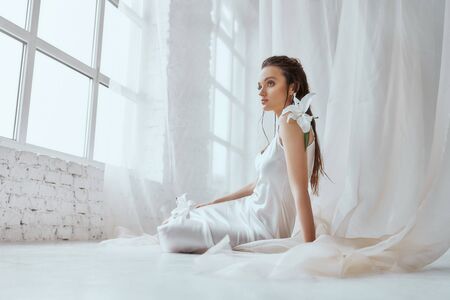 Side view of young beautiful brunette woman sitting on floor and holding lily flower on shoulder. Portrait of girl with wet hair posing on white background and looking out window. Concept of beauty.の写真素材