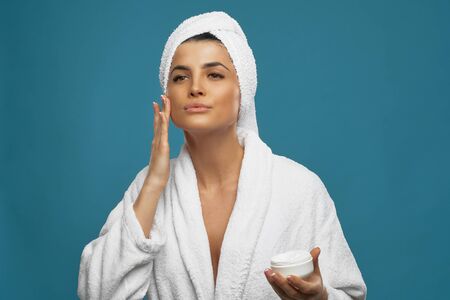 Front view of young woman in bathrobe and towel on head using cream for hydrating and rejuvenating...