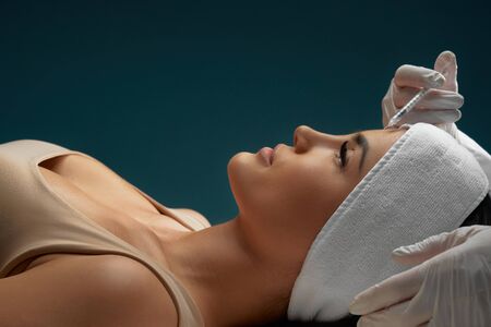 Close up of cosmetic botox injection in female forehead. Cosmetologist using syringe with liquid while brunette patient in towel on head lying, isolated on dark blue. Concept of cosmetology, beauty.の写真素材