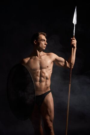 Front view of young athletic warrior with perfect body holding spear and shield. Crop of muscular bodubuilder posing with cold weapon on black studio background. Concept of warrior, strength.の写真素材