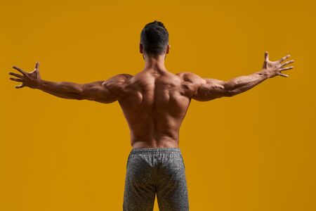Back view of shirtless bodybuilder with perfect biceps spreading muscular arms. Strong young man showing well trained body. Isolated on yellow studio background. Concept of strength and muscles.の写真素材