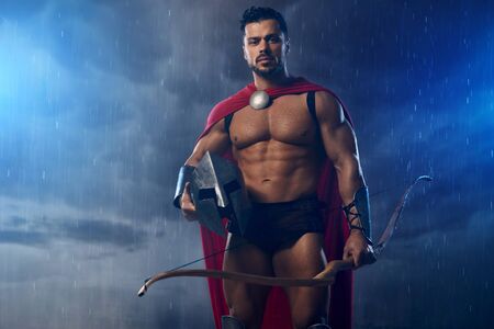 Front view of muscular bearded spartan wearing red cloak, holding bow and iron helmet while raining outdoors. Portrait of wet handsome man posing with weapon, looking at camera in overcast weather.の写真素材