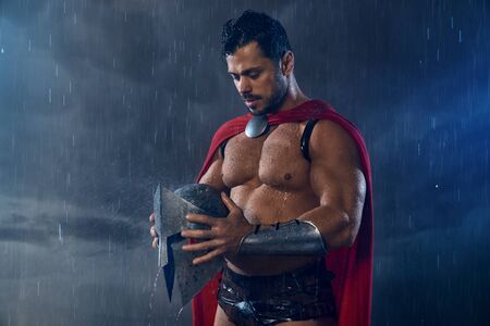 Portrait of muscular bearded spartan wearing red cloak and holding helmet while raining. Portrait of...