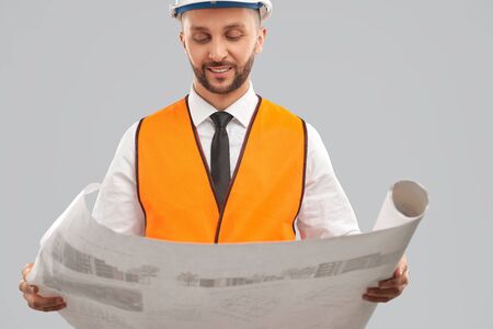 Crop of young bearded architect in orange reflective vest and helmet holding big map. Smiling...