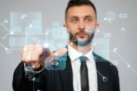 Front view of young serious businessman in black suit in office. Selective focus of digital tactile charts screen, bearded man clicking virtual icon. Concept of high technologies, digitalization.の写真素材