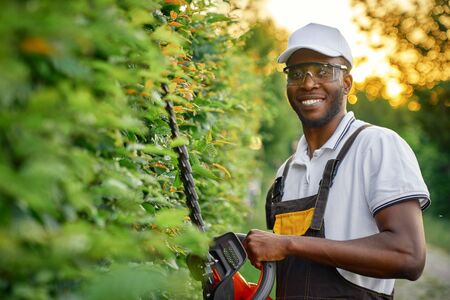 Happy african man wearing special clothing and protective glasses cutting bushes with petrol hedge...