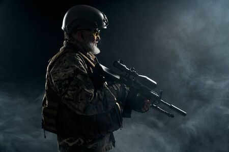 Side view of old military officer holding sniper rifle and looking away. Portrait of bearded veteran...