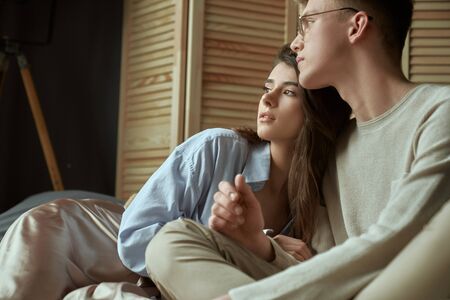 Front view of sweet happy young couple sitting and hugging on bed at home. Crop of young stylish brunette girl relaxing in arms of boyfriend in glasses. Concept of love, relationship.の写真素材