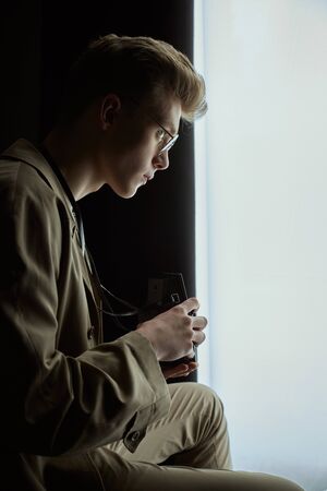 Side view of young blonde male photographer sitting in bedroom near window with black curtains. Crop...