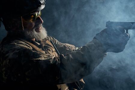 Side view of old military officer holding small gun and aiming. Portrait of bearded veteran in...