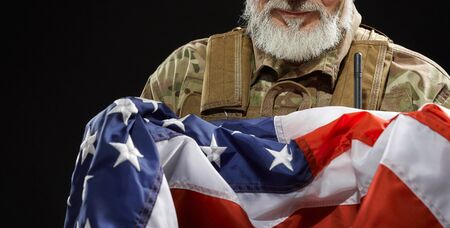 Close up of bearded american military veteran holding flag in arms. Portrait of incognito old proud male officer in camouflage uniform posing in dark atmosphere. Concept of military, patriotism.の写真素材