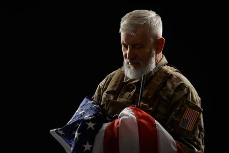 Front view of bearded american military veteran holding flag in arms. Portrait of old proud male officer in camouflage uniform posing in dark atmosphere. Concept of military, patriotism.の写真素材