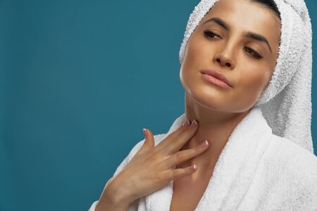 Front view of young woman in bathrobe and towel on head using cream for hydrating of soft neck skin. Portrait of pretty female after shower, isolated on blue. Concept of beauty, skincare.の写真素材