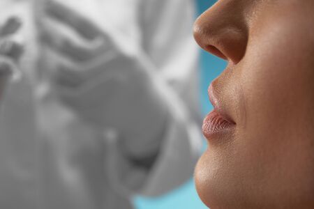 Close up of female face and incognito doctor in gloves and white coat behind. Crop of caucasian woman waiting for procedure, isolated on blue. Concept of cosmetology, beauty.の写真素材