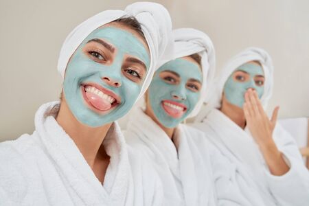 Close up of smiling group of girls with cosmetic masks, in towels on heads and bathrobes taking...