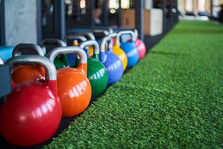 Close up of colorful kettle bells on floor near fake green grass. Selective focus of heavy steel equipment in gym, daylight. Concept of sport, weightlifting.の写真素材