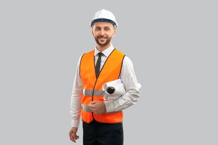 Front view of young architect in orange reflective vest and helmet holding big paper rolls. Smiling...