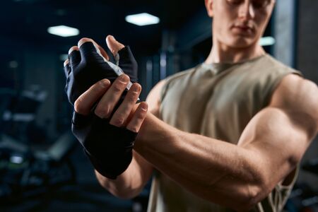 Crop of handsome bodybuilder in sportswear and gloves stretching upper body in sports club. Side view of muscular sportsman in gloves posing in empty gym, selective focus. Concept of bodybuilding.の写真素材