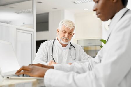 Therapists with stethoscopes on necks posing on reception in hospital. African male doctor using laptop, selective focus of caucasian eldery doctor holding pen and writing in folder on table.の写真素材