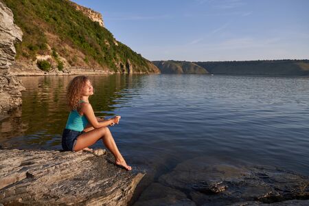 Romantic, pretty girl with long curly hair sitting bare foot on shore of lake. Beautiful, seductive...