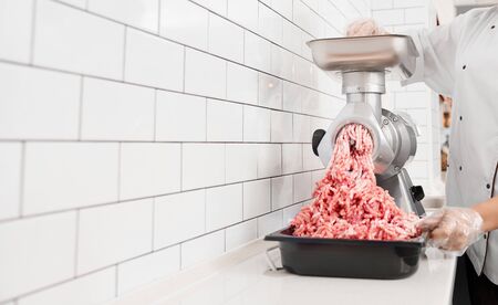 Crop of incognito woman with curly hair putting small pieces in meat grinder and grinds them into mincemeat. View of female working in butchery, preparing meat for selling in shop.の写真素材