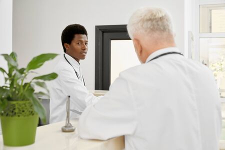 Selective focus of younf african doctor with stethoscope on neck standing behind reception and looking at coworker. Back view of incognito eldery therapist leaning on stand with plants.の写真素材
