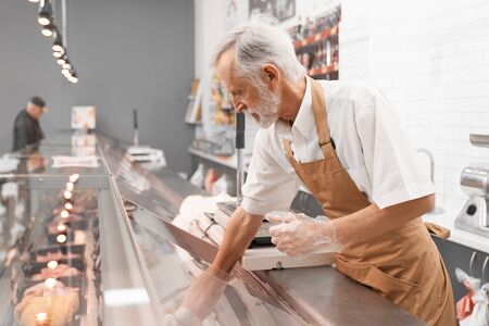 Side view of senior male butcher putting out of glass counter plate with sliced fresh steaks. Raw meat pieces in refrigerator with price tags ready for sale in meat department. Concept of food.