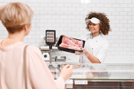 Young smiling woman with curly hair demonstrating to lady raw pork. Cheerful girl behind store counter with big assortment showing meat in bowl from refrigerator, supermarket.の写真素材