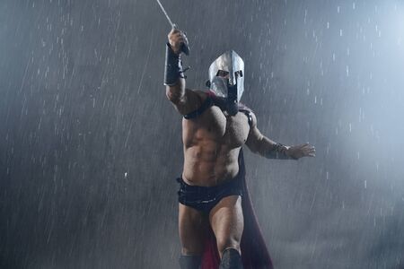 Front view of wet roman gladiator in iron helmet and red cloak attacking with sword. Muscular screaming shirtless spartan in red cloak and armor running during fight in rainy bad weather.の写真素材