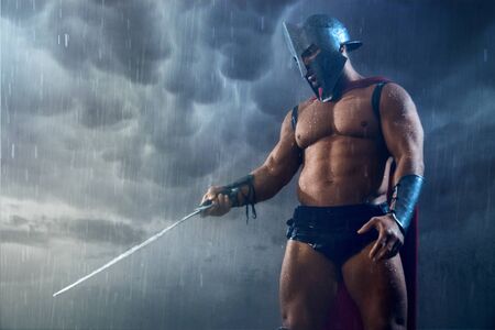 Front view of wet roman gladiator in iron helmet pointing sword down. Muscular shirtless spartan in red cloak and armor looking at raindrops on blade during fight in rainy bad weather, grey sky.の写真素材