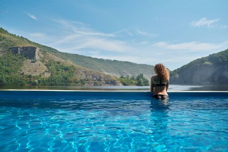 Beautiful, sexy girl wearing in black swimsuit standing in blue water leaning on edge of pool. Seductive lady with long curly hair admiring landscape and resting.の写真素材