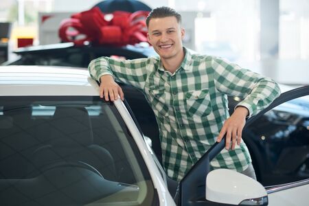 Portrait of handsome smiling man posing, leaning on new car with door open in dealership, looking at...