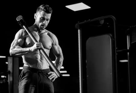 Front view of muscular man holding big sledgehammer on shoulder. Monochrome portrait of shirtless caucasian bodybuilder building muscles in gym in dark atmosphere. Concept of bodybuilding, sport.の写真素材