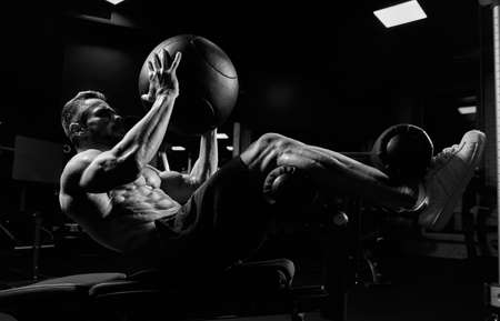 Side view of muscular man training abs with ball on bench in gym. Monochrome portrait of handsome adult sportsman building core muscles, doing crunches in dark atmosphere. Concept of sport, fitness.の写真素材