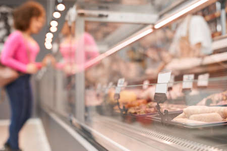 Side view of fasionable lady with curly hair looking and choosing fresh meat in store. Selective focus of store counter with big assortment of raw meat products in supermarket.の写真素材