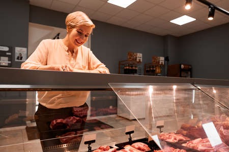 Front view of fasionable smiling lady looking and choosing fresh meat in store. Cheerful woman standing near store counter with big assortment of raw meat products in supermarket.の写真素材