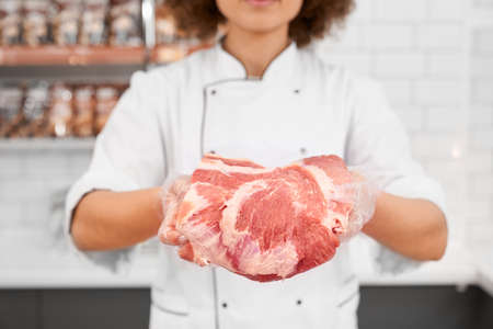 Selective focus of fresh red raw piece of meat in hands of young incognito woman with curly hair. Crop of girl behind counter of store showing pork from refrigerator in supermarket.の写真素材