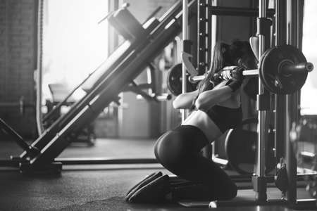 Side view of brunette female bodybuilder using wireless headphones and doing squats on knees in multipower smith machine. Woman in black outfit with fit body training in gym, black and white.の写真素材