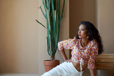 Portrait of mulatto girl with long black curly hair posing on floor near big cactus and wooden...