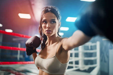 Close up of fit concentrated brunette woman wearing boxing gloves. Young attractive fighter with perfect make up posing and showing hit on boxing ring, colorful lights on background.の写真素材
