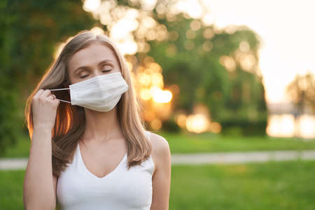 Portrait of young attractive woman with eyes closed taking off white protective face mask. Pretty female model posing in summer park, beautiful sunset on background. Pandemic concept.の写真素材