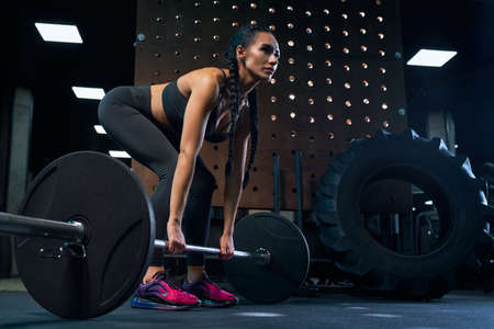 Side view of stunning female bodybuilder with long braids doing deadlift using barbell. Srtong woman with perfect muscular body in sportswear training legs in gym in dark atmosphere, full length.の写真素材