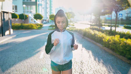 Front view of young cjarming woman wearing sports shorts and hood on head practicing morning jogging, looking down. Female runner practicing cardio workout in streerts, multistorey buildings around.の写真素材