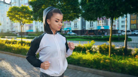 Young beautiful woman wearing jacket with hood on head jogging in sunny summer morning in city streets. Stunning female runner doing cardio workout outdoors, multistorey buildings around.の写真素材