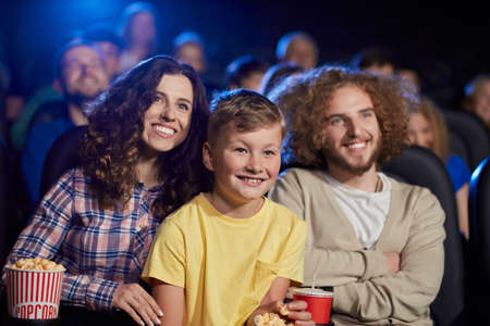 Selective focus of young parents with curly hair enjoying time with son holding sparkling drink, audience on background. Happy family sitting in cinema, kid on knees, watching funny cartoon.の写真素材