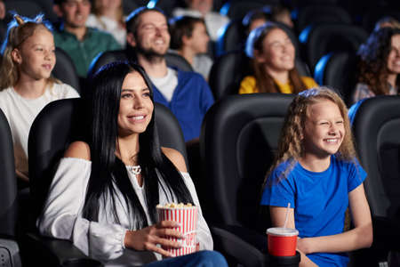 Side view of stunning brunette girl holding popcorn, watching comedy with younger sister in cinema,...