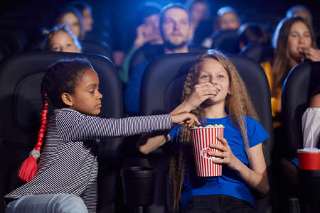 Kids enjoying time together, young multiracial audience on background. Happy friends sitting in cinema, watching funny cartoon, caucasian girl eating and sharing popcorn with african female friend.の写真素材