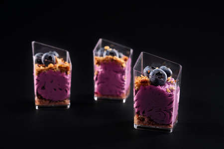 Front view of delicious sweet muesli dessert in three small glasses in row. Tasty parfait with granola, blueberries on top and bright purple whipped cream isolated on black background.の写真素材