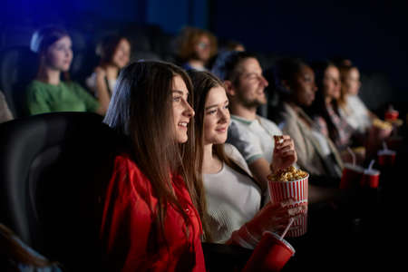 Selective focus of young female teen friends watching movie in cinema, sitting in comfortable seats. Side view of smiling caucasian girl enjoying film with group of people. Entertainment concept.の写真素材
