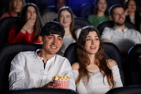 Selective focus of young man laughing, holding popcorn while watching comedy in cinema. Front view of caucasian man and pretty woman enjoying funny film. Concept of entertainment.の写真素材
